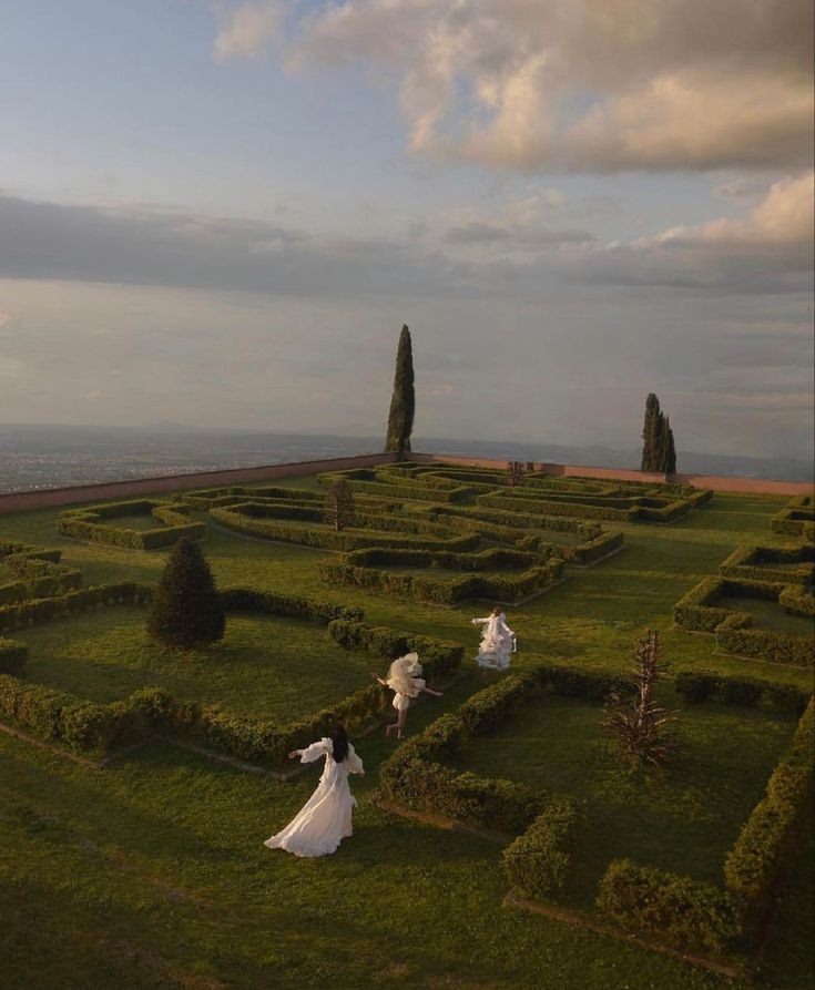 two women in white dresses are walking through an elaborate mazed area with trees and bushes