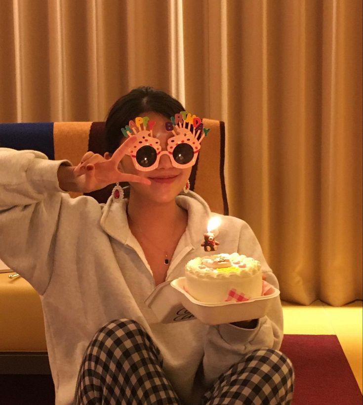 a woman sitting in a chair holding a plate with a cake on it while wearing pink glasses