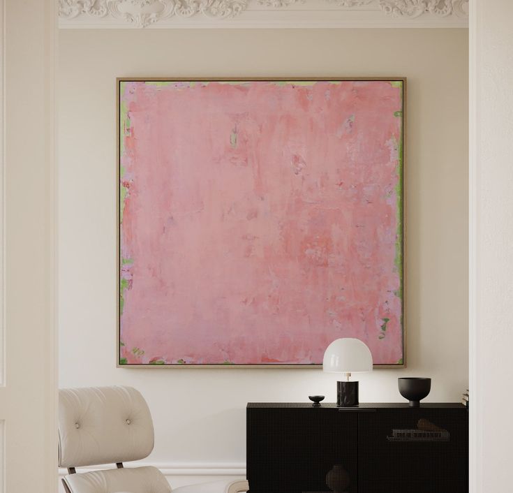 a large pink painting hangs above a white chair in front of a black cabinet and lamp