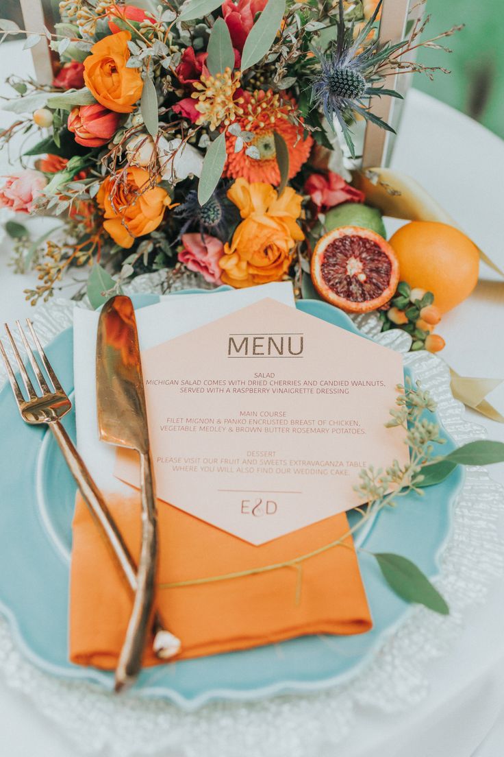 an orange and blue table setting with silverware, napkins, and flowers on it