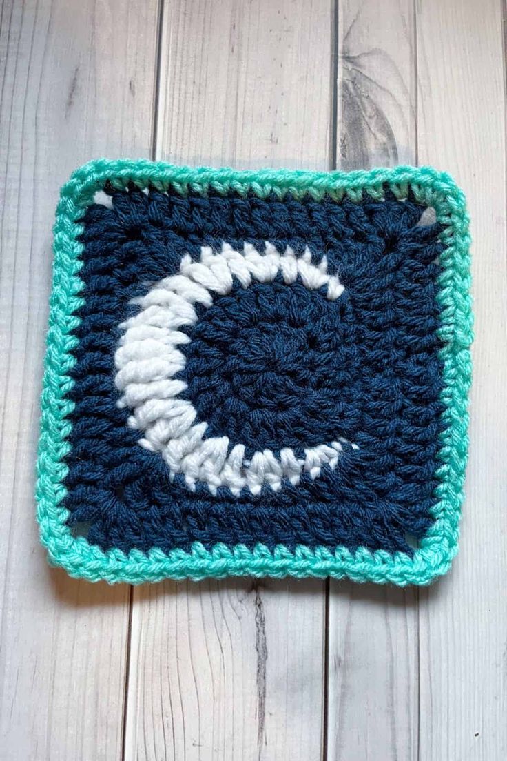 a crocheted square with a white and blue eyeball on the center is sitting on a wooden surface