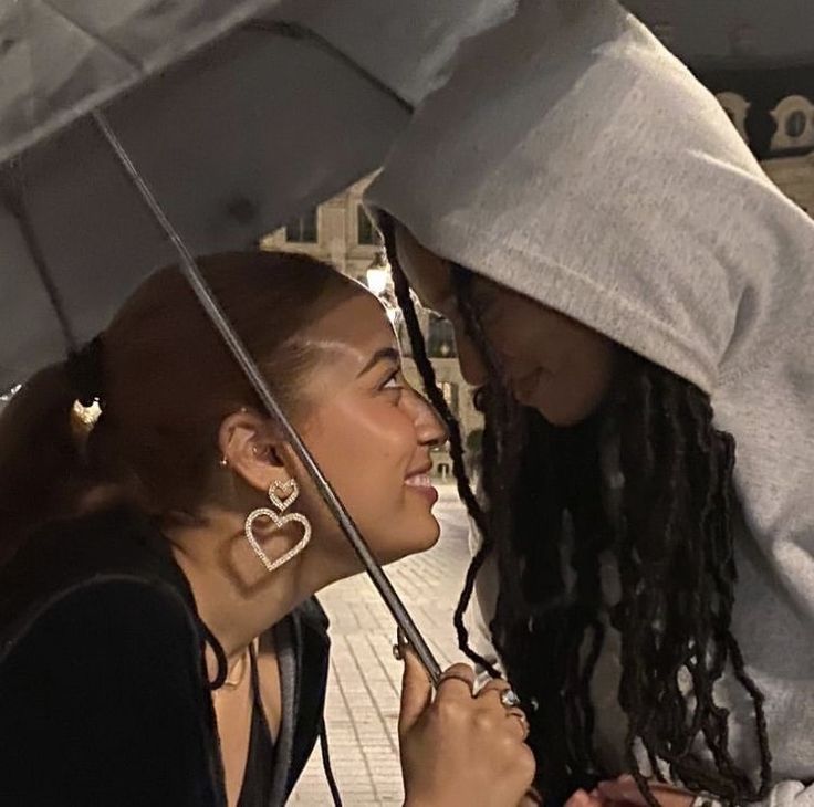 two young women standing under an umbrella in the rain, one holding her earring