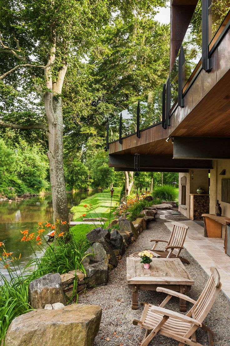 an outdoor patio with wooden chairs and tables next to a river in the background, surrounded by greenery