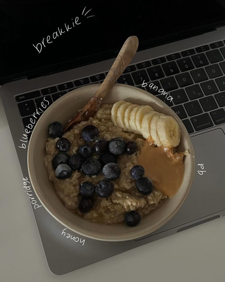 a bowl of oatmeal with bananas and blueberries in front of a laptop