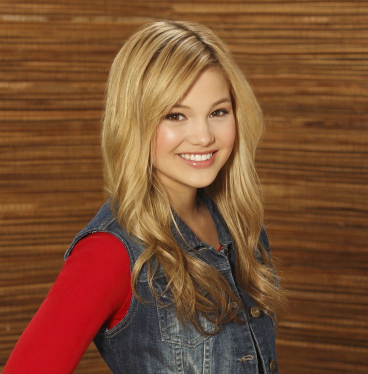 a woman with long blonde hair wearing a jean jacket and red shirt smiling at the camera