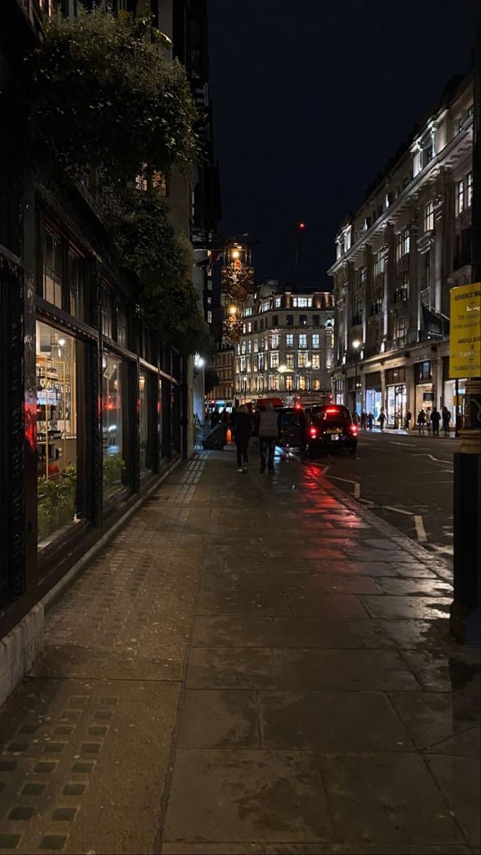 a city street at night with cars parked on the side walk and people walking down the sidewalk