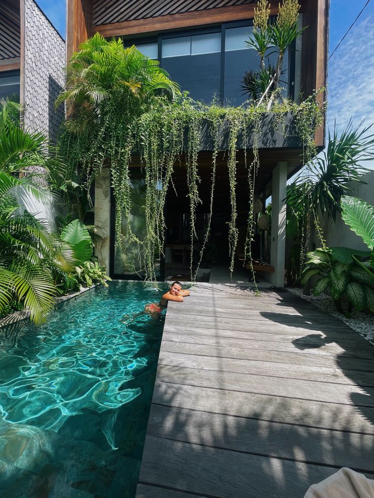 an outdoor swimming pool surrounded by greenery and wooden walkway leading to a large house