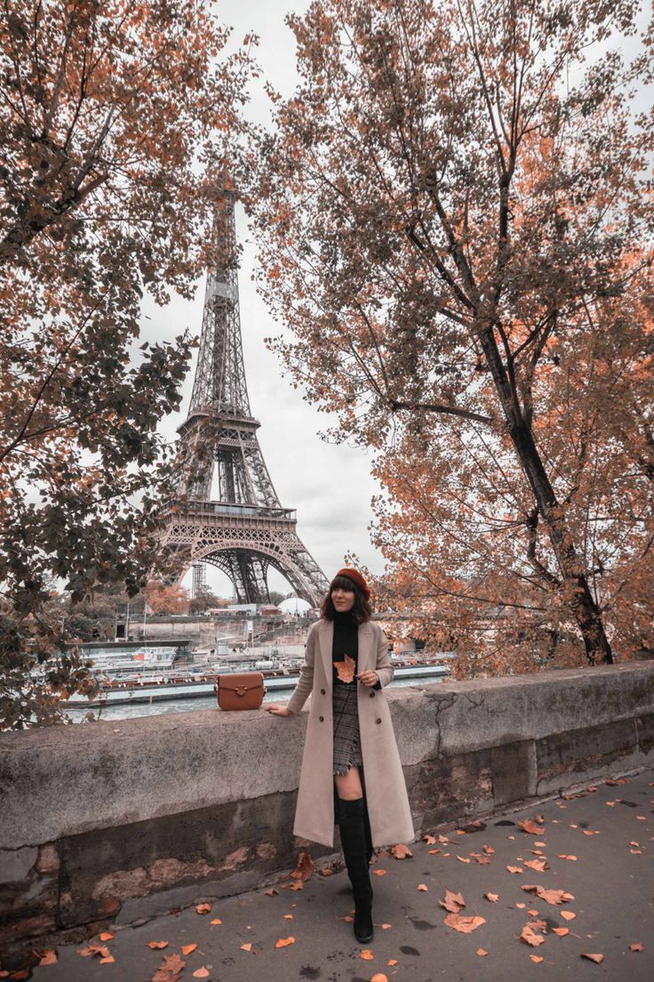 a woman standing in front of the eiffel tower with autumn leaves on the ground