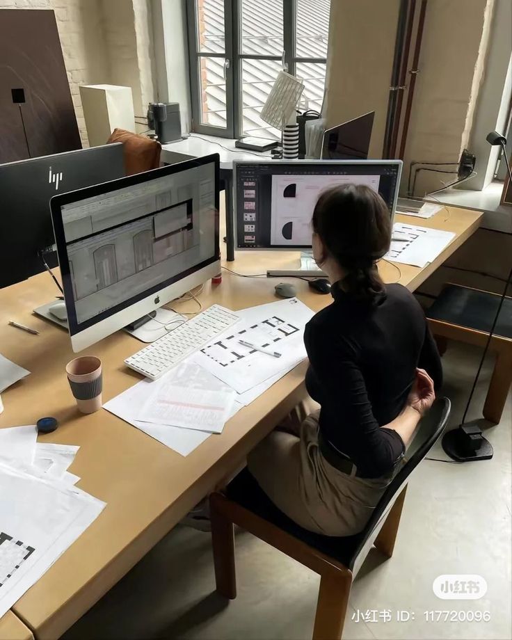 a woman sitting at a desk in front of two computer monitors with papers on it