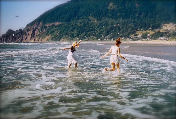 two young women are playing in the water at the edge of the beach, with mountains in the background