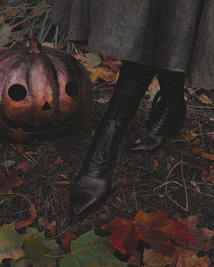 a woman in black boots standing next to a pumpkin on the ground with leaves around her