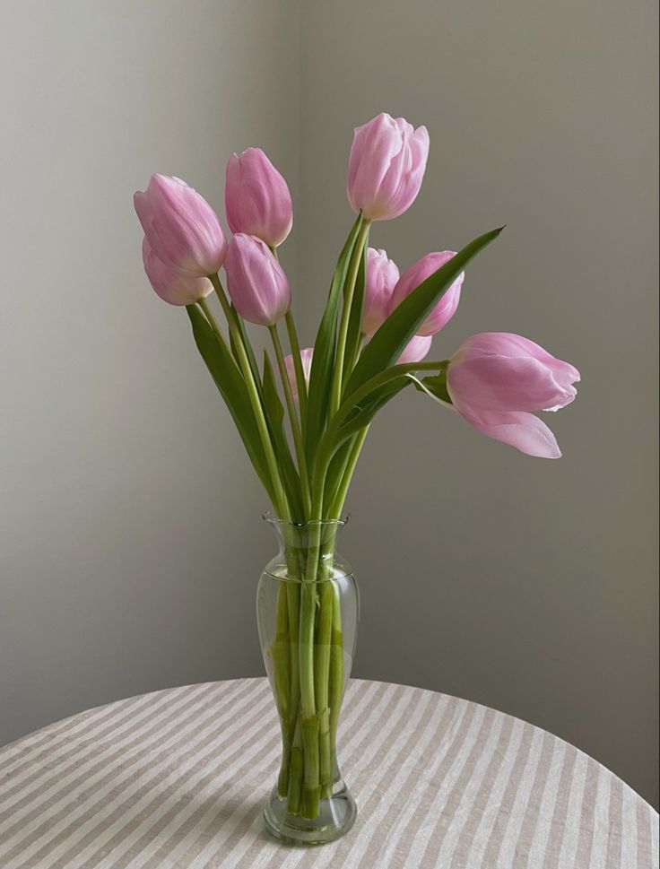 pink tulips are in a clear vase on a striped tablecloth with a gray wall behind it