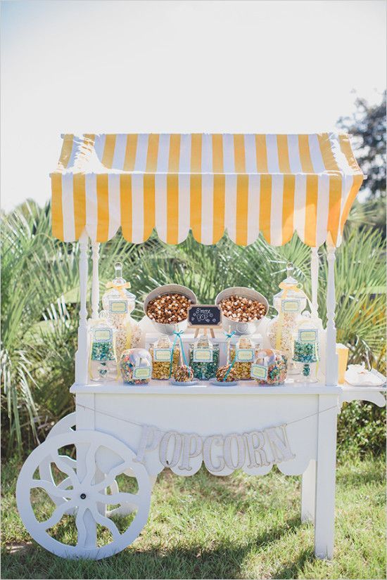 an old fashioned popcorn cart is set up in the grass for a wedding reception with yellow and white striped awning