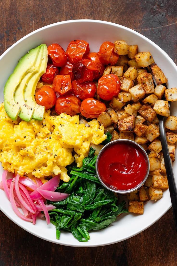 a white plate topped with eggs, tomatoes and other foods next to a bowl of ketchup