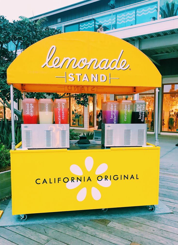 a yellow lemonade stand with cups on it in front of a shopping center building