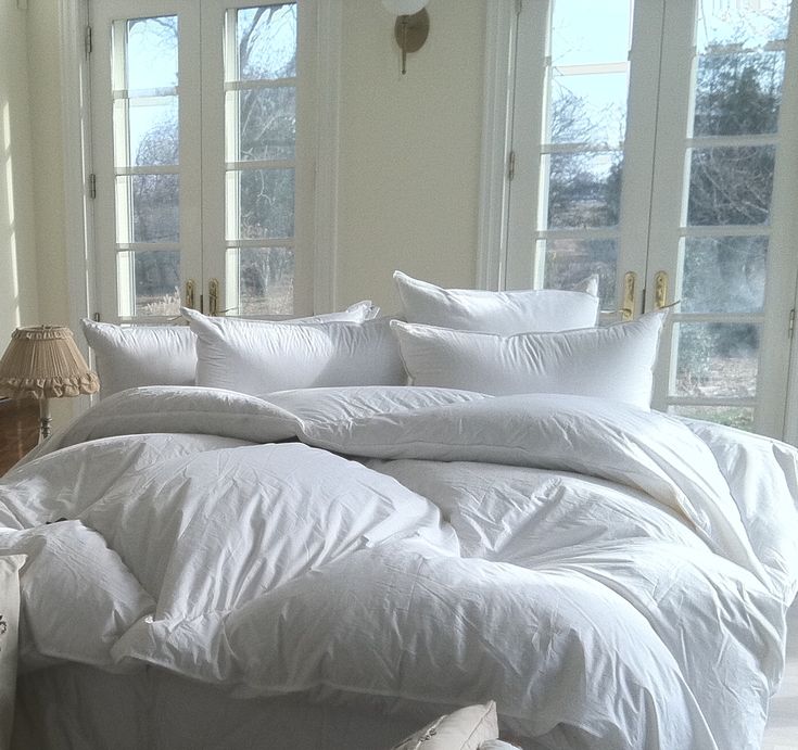 a bed with white sheets and pillows in a room next to two large windows on either side of the bed