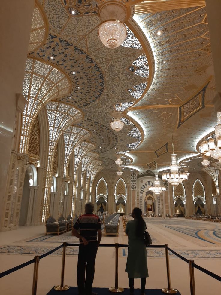 two people standing in the middle of a large hall with chandeliers hanging from the ceiling