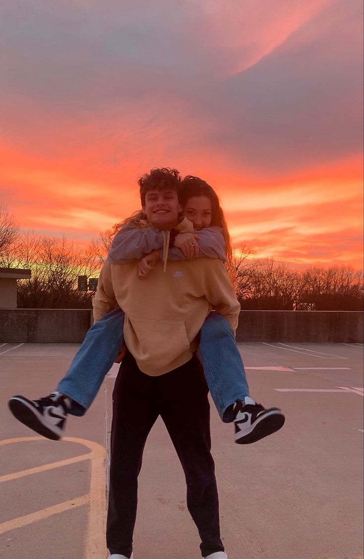 two people standing in a parking lot with the sun setting behind them and one person holding his arms around the other