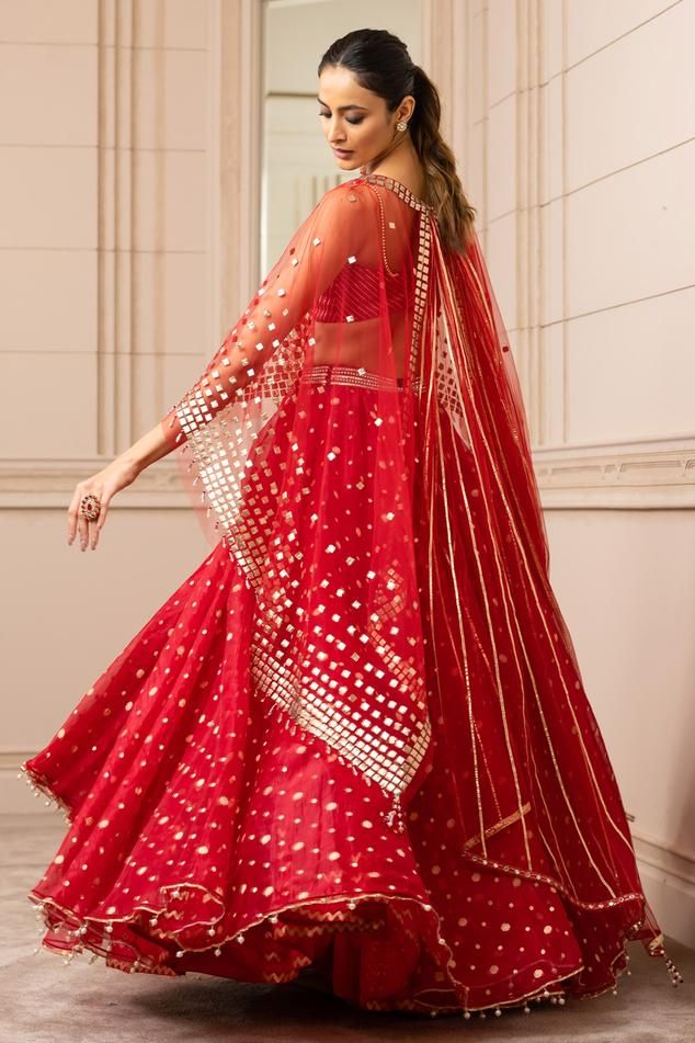 Red asymmetric organza brocade lehenga with an under-layer of chevron borders. Paired with a textured bustier with fringe embellishment. Comes with a sheer cape with square-cut gota embroidery and gota-lined wings extended in the back.
Component: 3
Pattern: Embroidered
Type Of Work: Geometric
Neckline: Cape: Round
Sleeve Type: Cape: Draped Sleeves
Fabric: Silk organza brocade
Color: Red
Other Details: 
Draped lehenga
Tassel detail
Gota work
Asymmetric cape
Occasion: Destination Wedding - Aza Fas Red Floor-length Choli With Gota Work, Floor-length Red Choli With Gota Work, Red Organza Dress For Navratri, Red Cutdana Choli For Party, Party Red Choli With Cutdana Details, Red Sharara With Sheer Dupatta In Chanderi, Red Saree With Sheer Dupatta For Reception, Red Organza Floor-length Saree, Floor-length Red Organza Saree