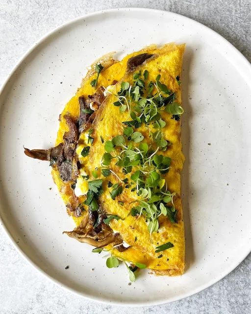 a white plate topped with an omelet covered in greens and mushrooms on top of a table