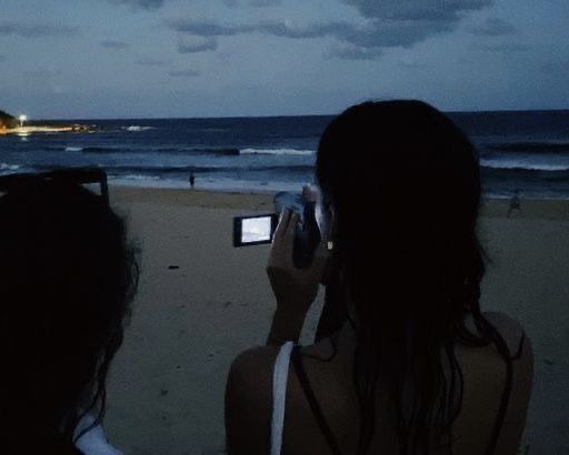 two people are taking pictures on their cell phones at the beach as the sun goes down