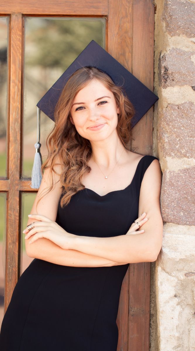 a young woman wearing a graduation cap and gown leaning against a wall with her arms crossed