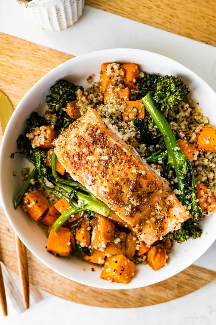 a white bowl filled with salmon, broccoli and carrots on top of a wooden cutting board