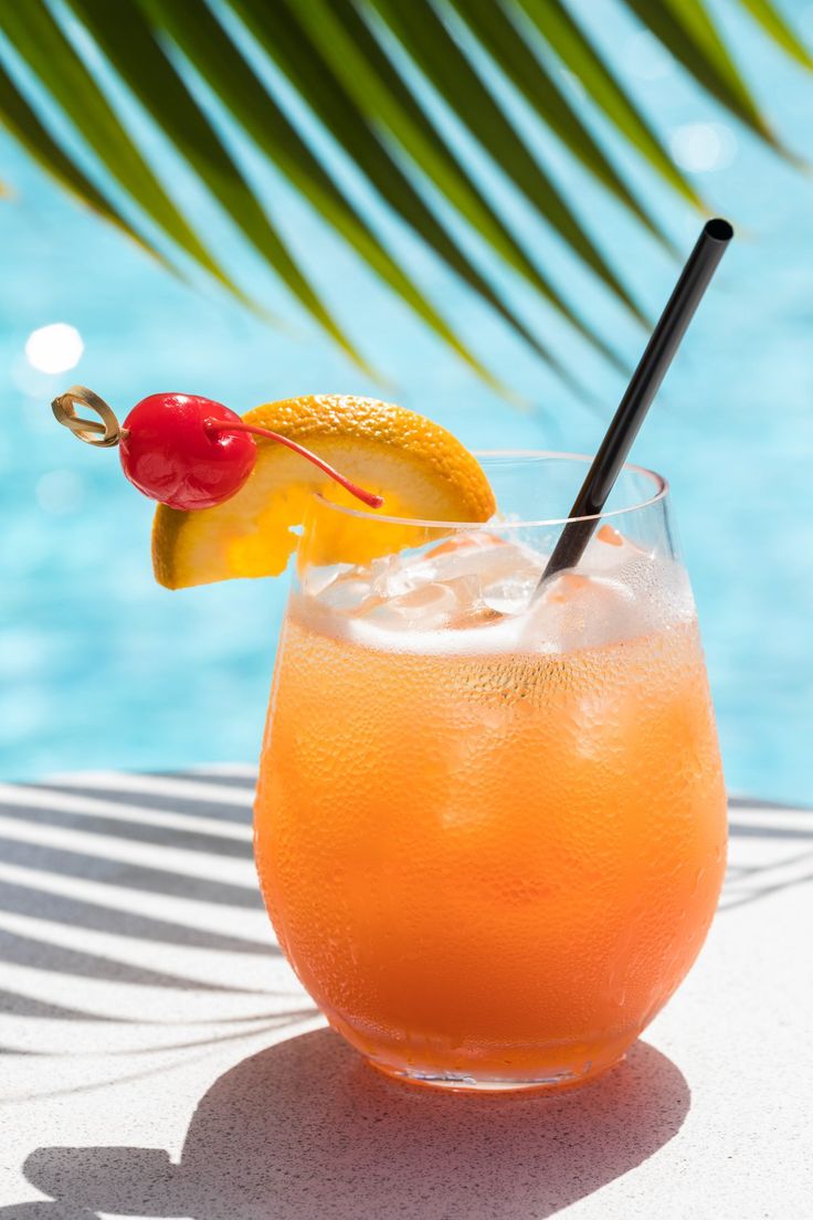 an orange drink sitting on top of a table next to a swimming pool with a cherry garnish