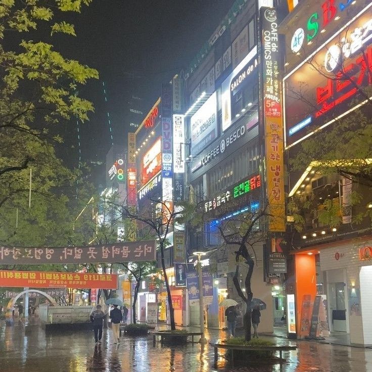 people are walking in the rain on a city street at night with neon signs and trees