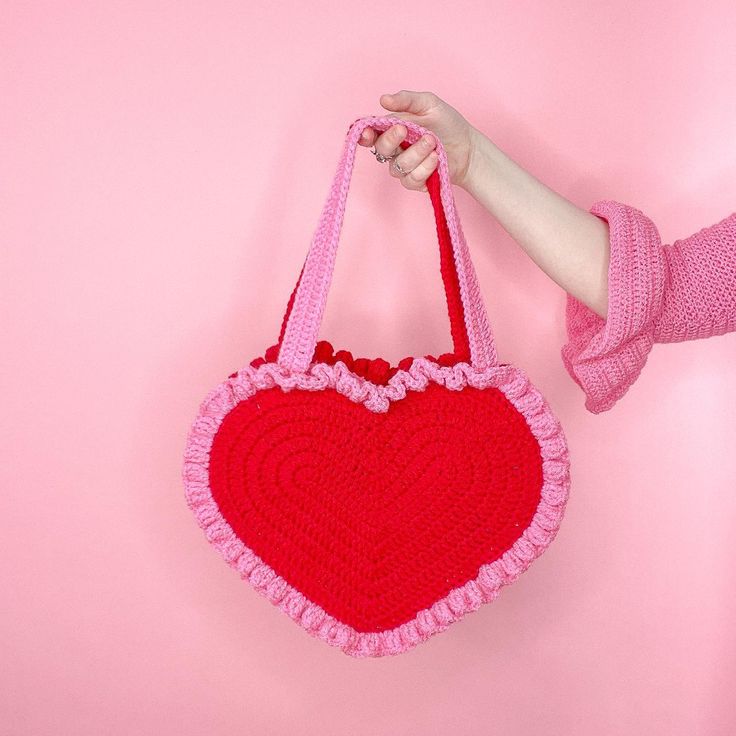 a crocheted heart shaped purse hanging from a hook on a pink wall, with someone's hand holding it