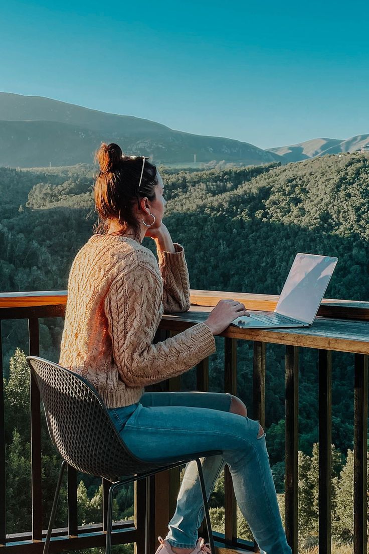 a woman sitting at a table with a laptop on her lap looking out over the mountains