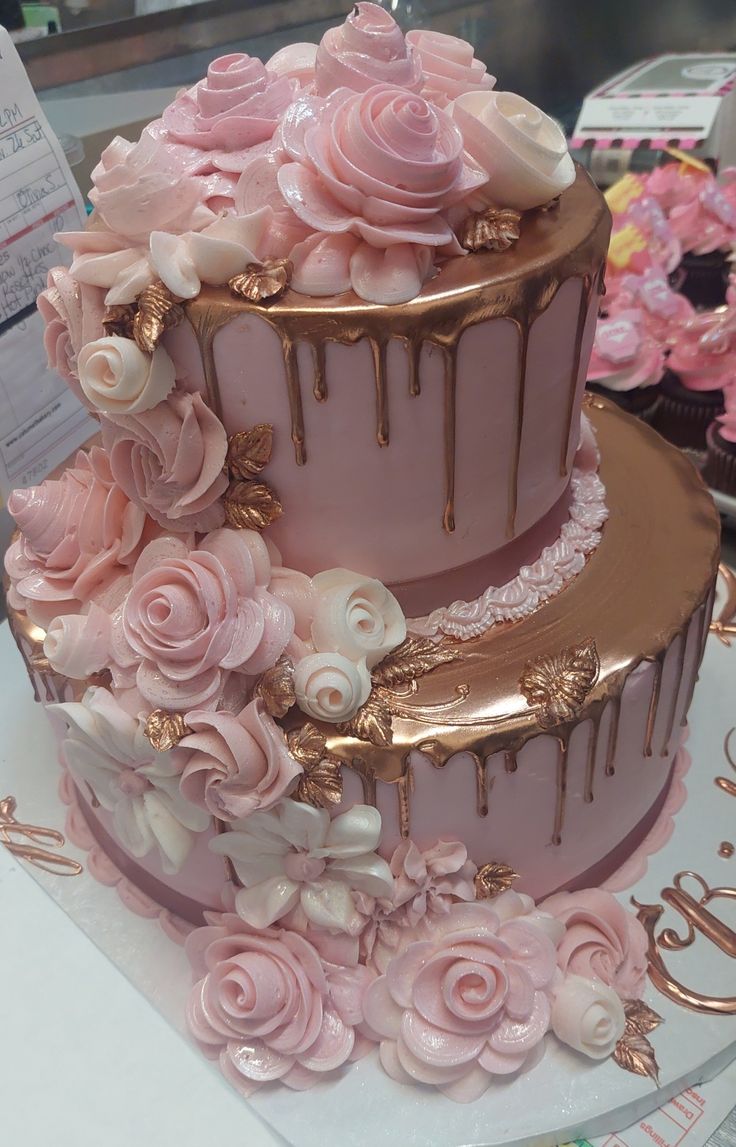 a three tiered cake with pink flowers and gold trimmings on display at a bakery