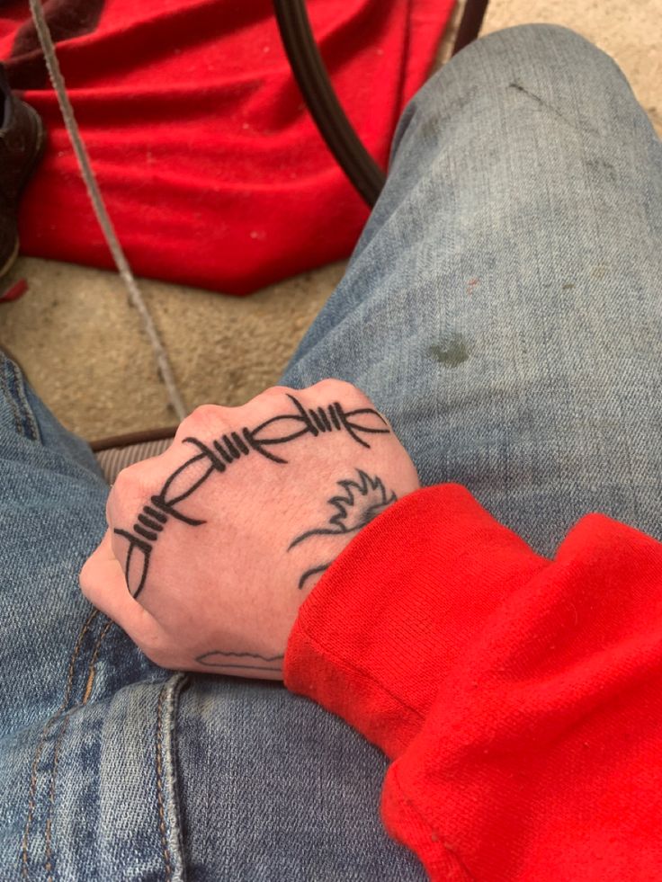 a person with a tattoo on their wrist sitting next to a red bag and wearing blue jeans