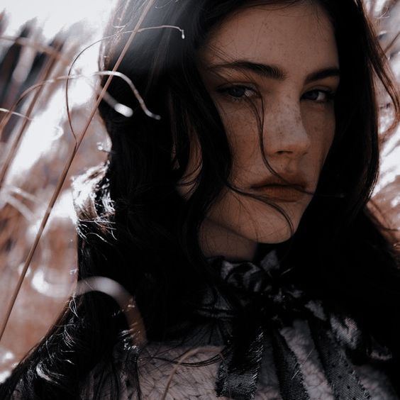 a woman with long black hair standing in front of some tall grass and looking off into the distance