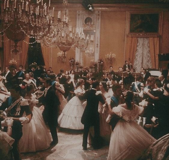 a group of people dressed in formal wear dance around a chandelier filled room