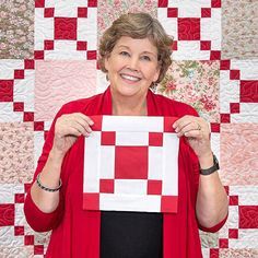 a woman holding up a piece of paper with red and white squares on it in front of a quilt