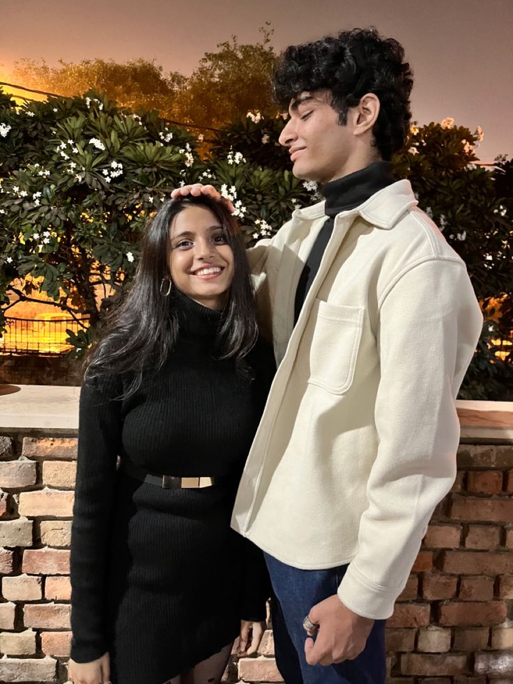 a man and woman standing next to each other in front of a brick wall at night