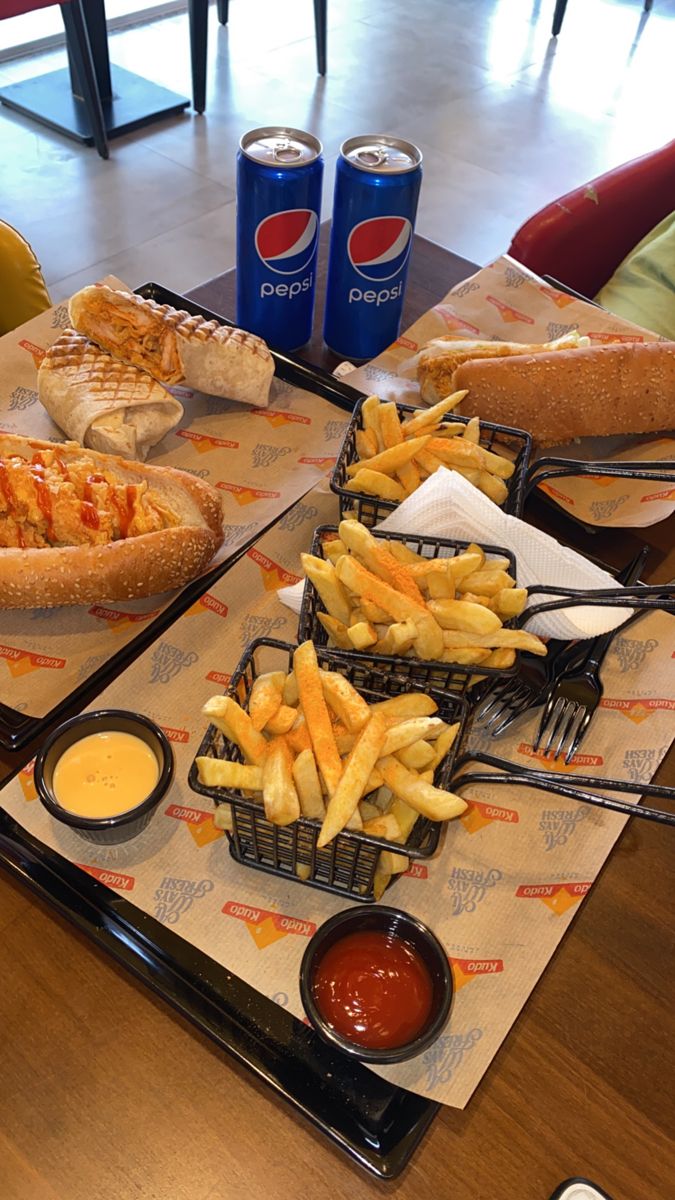 two trays filled with sandwiches and french fries on top of a table next to soda cans