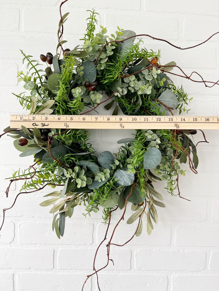 a green wreath hanging on a white brick wall next to a ruler with greenery