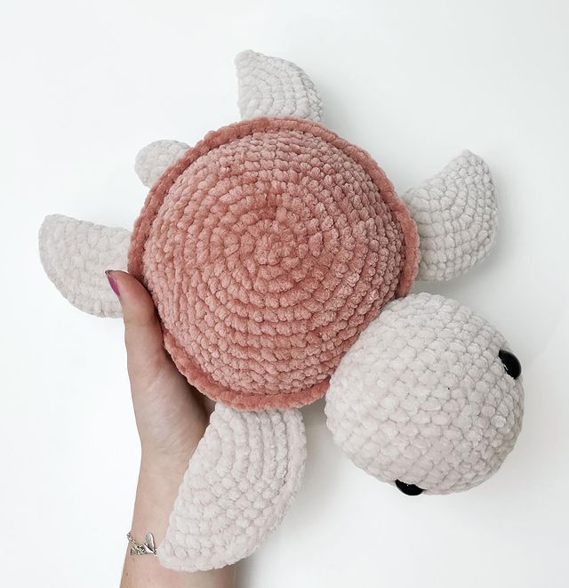 a hand holding a pink and white crocheted turtle toy with its head on it's back