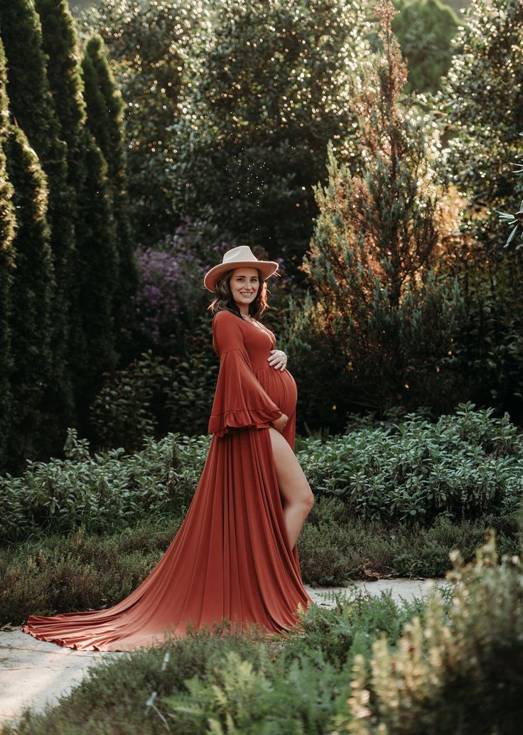 a pregnant woman wearing a long red dress and hat poses for a photo in the garden