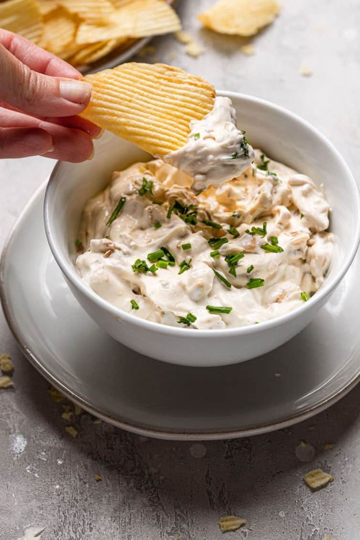 a hand dipping a cracker into a bowl of dip with chips on the side