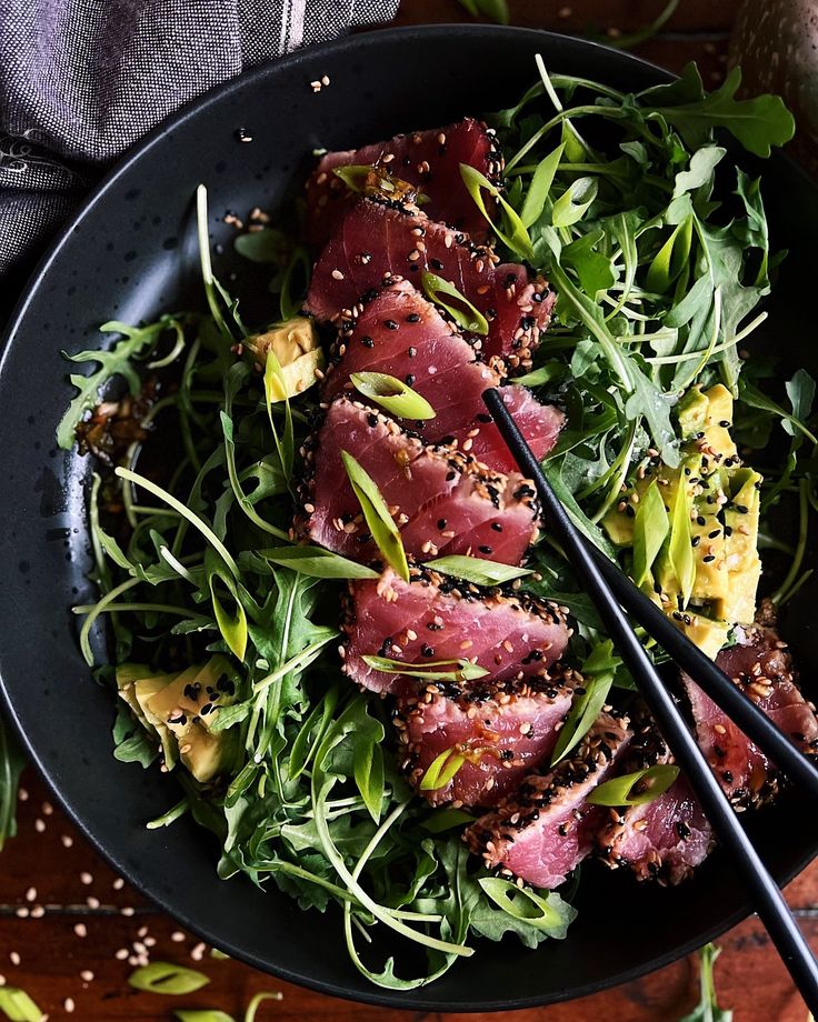 a black bowl filled with meat and greens next to chopsticks on top of a wooden table