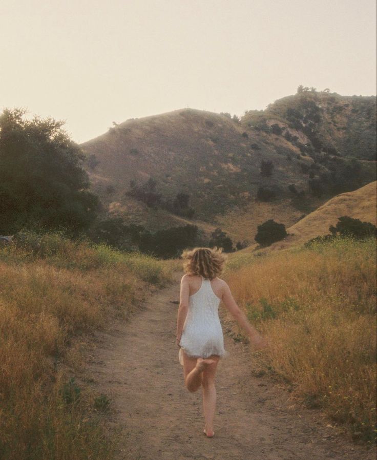 a woman walking down a dirt road in the middle of a field with hills behind her