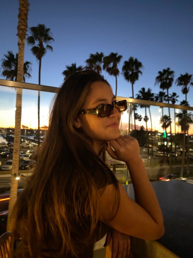 a woman wearing sunglasses standing next to a palm tree at night with the sun setting in the background