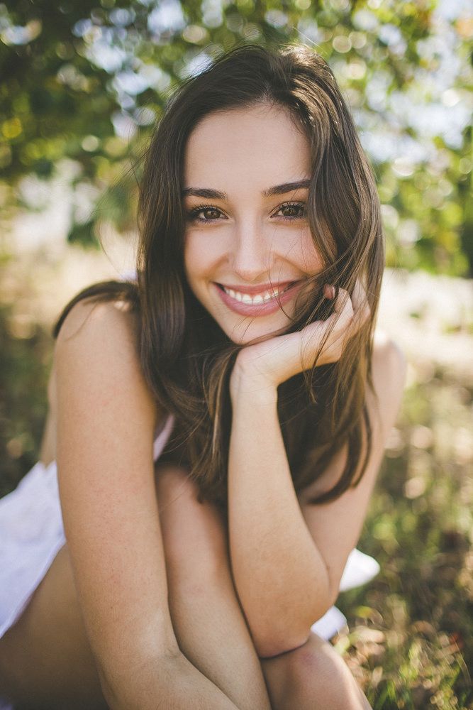 a beautiful young woman laying on the ground in front of trees smiling at the camera