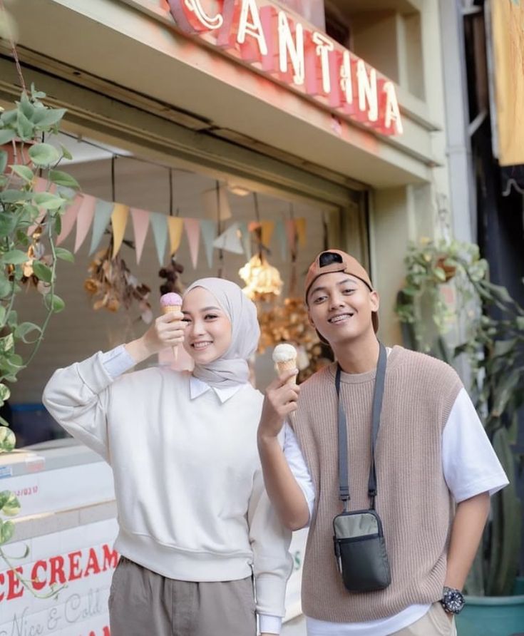 two people standing in front of a store holding up ice cream cones and looking at the camera