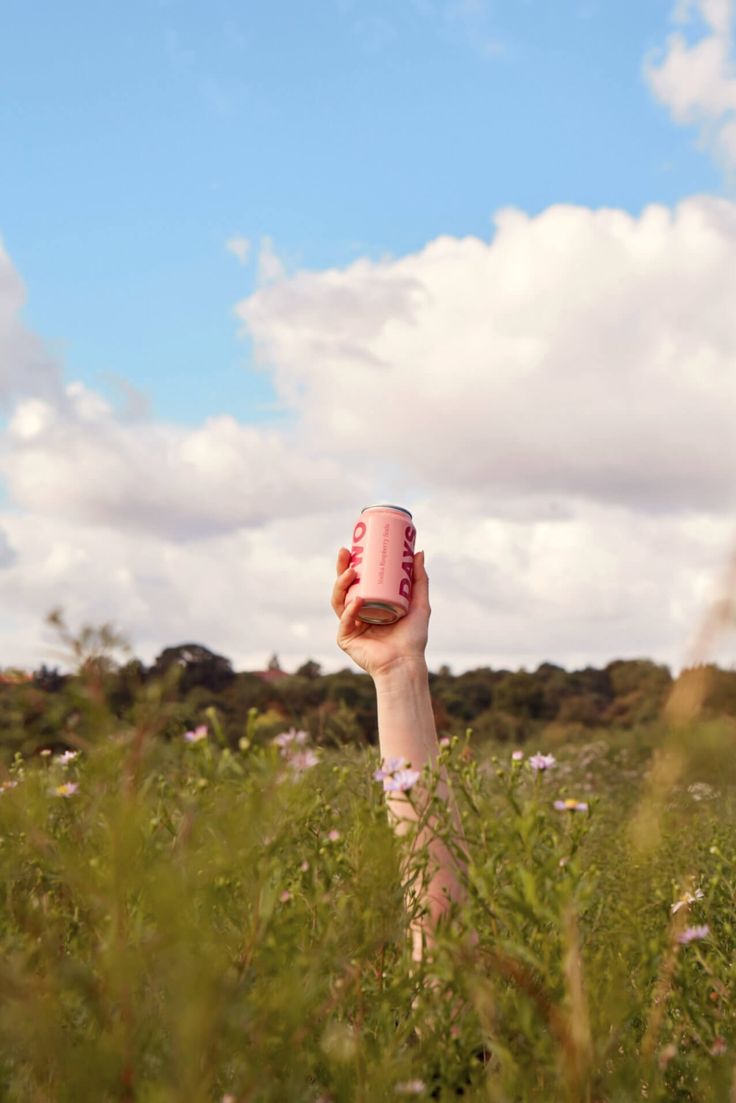 a person holding up a pink cup in the middle of a field with wildflowers