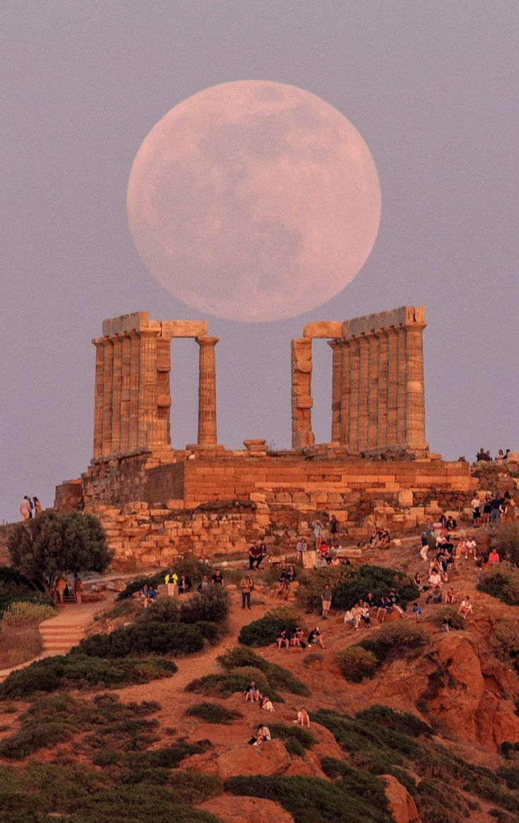 the full moon is setting over some ruins on top of a hill with people walking up to it