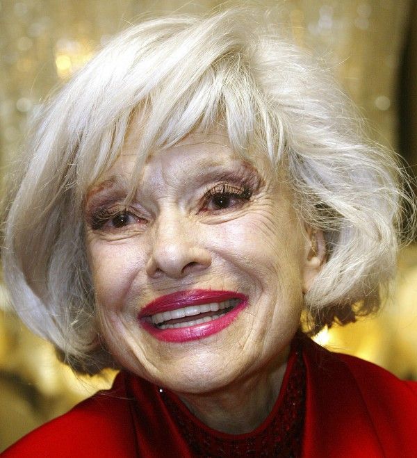 an older woman with white hair and bright red lipstick smiles at the camera while wearing a red jacket